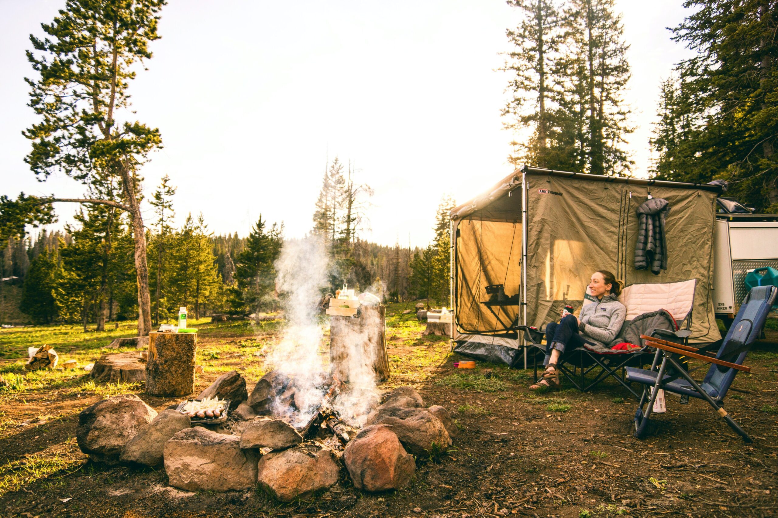 A campfire sits in the foreground, surrounded by small stones. A camper sits in a yellow tent in the background, not watching the fire. 